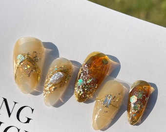 May include: Set of five false nails with a light brown base and gold glitter accents. Some nails have a clear top coat with gold glitter and a pearl accent. Others have a clear top coat with gold glitter and a small, clear crystal accent. The nails are arranged in a row on a white surface with the text "NG ORIGINAL" and "Make re" visible.
