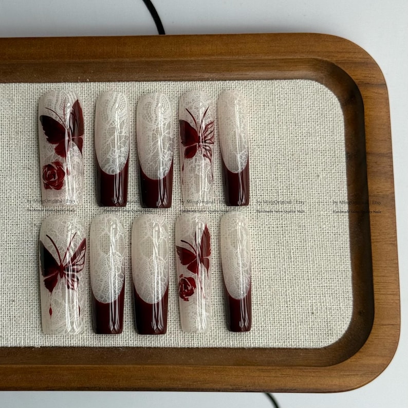 Pu&ograve; includere: Set di dieci unghie finte con un design in pizzo bianco, punte bordeaux e dettagli di farfalle e rose bordeaux. Le unghie sono presentate su un vassoio di legno con una base in lino. Il testo "by MingOriginal Etsy" &egrave; visibile.