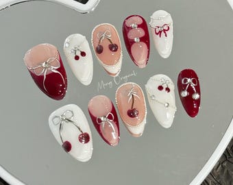 May include: A set of ten false nails with a cherry and bow design. The nails are white with red tips and feature a silver chain and a red cherry with a green stem. The nails are arranged on a white heart-shaped surface with the text "Mung Original" in black.