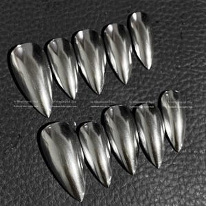 May include: A set of ten silver chrome-finish false nails. The nails are almond-shaped and have a reflective, metallic appearance. The nails are arranged on a dark surface.