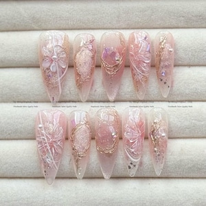 May include: A collection of ten long, almond-shaped artificial nails. Each nail has a light pink base with detailed designs, including floral accents, gold wire detailing, and sparkling glitter. The nails are embellished with small pearls and iridescent elements.