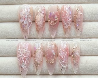 Könnte beinhalten: Ein Set aus zehn langen, mandelförmigen Kunstnägeln. Jeder Nagel hat eine hellrosa Basis mit aufwendigen Designs, darunter florale Akzente, goldfarbene Drahtdetails und glitzernde Glitzer. Die Nägel sind mit kleinen Perlen und irisierenden Elementen verziert.