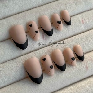 Può includere: Set di dieci unghie finte color nude con punte nere e dettagli dorati. Ogni unghia ha un piccolo design a forma di cuore nero.