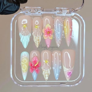 May include: A set of ten false nails in a clear plastic case. The nails are decorated with a variety of summer-themed designs, including seashells, flowers, and stars. The nails are in shades of pink, blue, and yellow.
