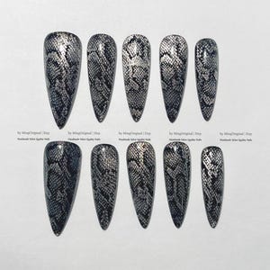 May include: A set of ten long, pointed false nails with a black and silver snakeskin pattern. The nails are arranged in two rows of five, showcasing the detailed design. Text at the bottom reads "by MingOriginal | Etsy Handmade Salon Quality Nails."