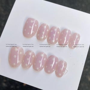 May include: A set of oval-shaped, light pink press-on nails with white polka dots. The nails have a glittery, iridescent finish at the base. The text "by MingOriginal | Etsy Handmade Salon Quality Nails" is printed on the card.