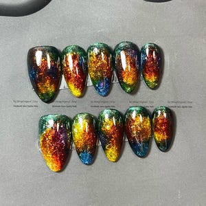 Pode incluir: Um conjunto de dez unhas postiças ovais com um acabamento brilhante e iridescente. As unhas fazem uma transição de um azul profundo nas pontas para tons vibrantes de verde, amarelo, laranja e vermelho, criando um efeito colorido e chamativo. As unhas estão dispostas em uma superfície cinza.