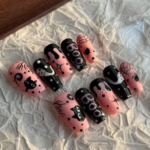 Könnte beinhalten: Ein Satz von zehn Sarg-förmigen Kunstnägeln. Die Nägel sind in Rosa und Schwarz bemalt und mit Halloween-Motiven versehen, darunter eine schwarze Katze, Spinnweben und das Wort "Boo".