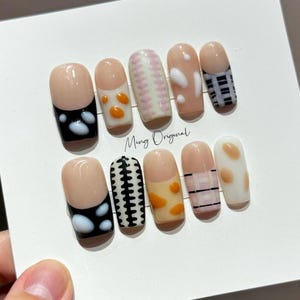 May include: A set of ten artificial nails with various designs. The nails feature a nude base with black, white, orange, and pink accents. Some nails have patterns like polka dots, plaid, and abstract shapes. The text "Ming Original" is visible.