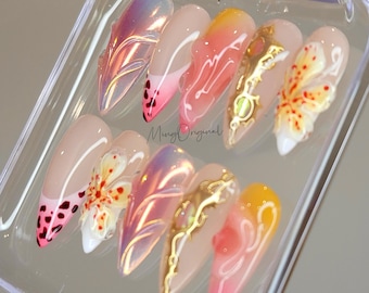 May include: A set of ten false nails with various designs. The nails are in a clear plastic case. Some nails have a pink and white ombre design, others have a butterfly design, and others have a floral design. Some nails have gold accents.