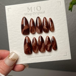May include: Set of ten brown and white marbled fake nails with a pointed oval shape. The nails are arranged in two rows of five on a white card with the text "MIO Ming Original" and "Luxury Handmade Nails".