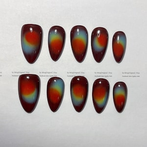 May include: A set of ten oval-shaped false nails. Each nail has a gradient design, transitioning from a deep burgundy at the base to a vibrant orange and then to a light blue at the tip. The nails are arranged on a white surface.
