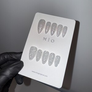 K&ouml;nnte beinhalten: Ein Set aus silbernen Glitzer-Press-On-N&auml;geln in verschiedenen Gr&ouml;&szlig;en und Formen, pr&auml;sentiert auf einer wei&szlig;en Karte. Die Karte zeigt den Text "M | O" und die Website "www.mingoriginal.com". Die N&auml;gel werden von einer behandschuhten Hand gehalten.