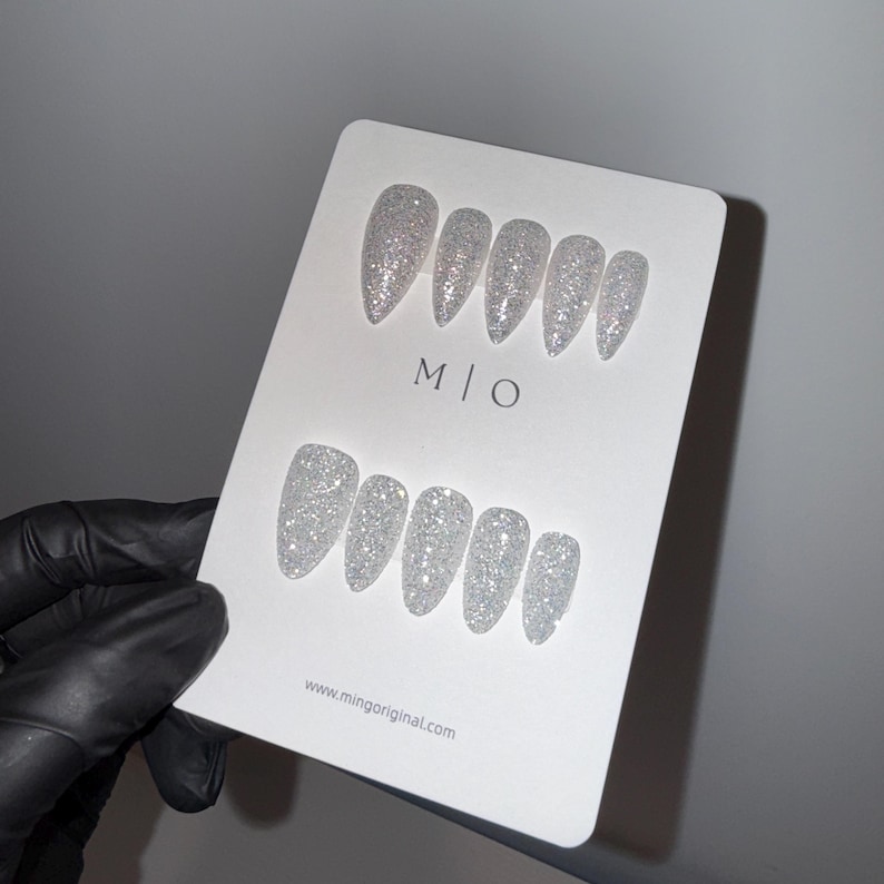 K&ouml;nnte beinhalten: Ein Set aus silbernen Glitzer-Press-On-N&auml;geln in verschiedenen Gr&ouml;&szlig;en und Formen, pr&auml;sentiert auf einer wei&szlig;en Karte. Die Karte zeigt den Text "M | O" und die Website "www.mingoriginal.com". Die N&auml;gel werden von einer behandschuhten Hand gehalten.