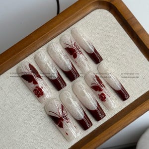 Pu&ograve; includere: Set di dieci unghie finte lunghe a forma di bara. Ogni unghia presenta un design in pizzo bianco su una base nude, con una punta bordeaux scuro. Alcune unghie hanno accenti di farfalle o rose bordeaux.