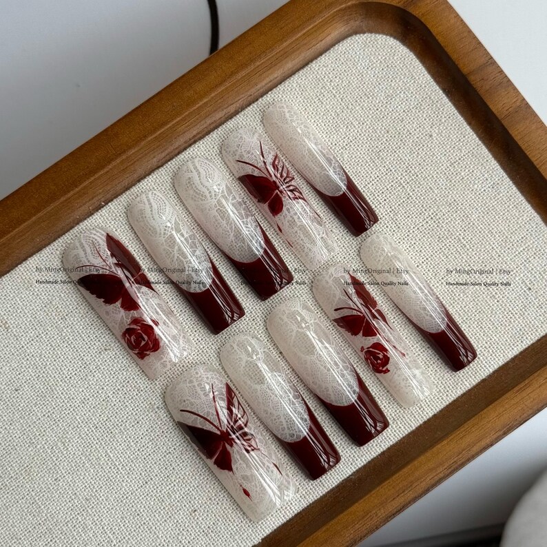 Pu&ograve; includere: Set di dieci unghie finte lunghe a forma di bara. Ogni unghia presenta un design in pizzo bianco su una base nude, con una punta bordeaux scuro. Alcune unghie hanno accenti di farfalle o rose bordeaux.