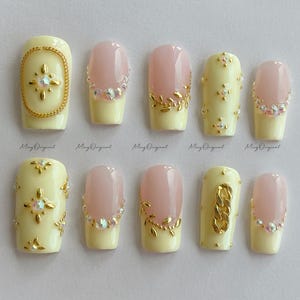May include: A set of ten artificial nails in various designs. Some nails are pale yellow with gold chain or floral accents and rhinestones. Others are pink with a pale yellow base and rhinestone or gold leaf accents. The brand name "Ming Original" is printed on the bottom.