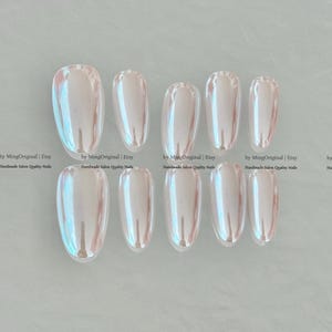 May include: Ten iridescent, oval-shaped false nails arranged in two rows. The nails have a pearlescent finish with hints of pink and blue. The text "by MingOriginal | Etsy Handmade Salon Quality Nails" is visible.
