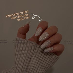 K&ouml;nnte beinhalten: Nahaufnahme einer Hand mit langen, stilettof&ouml;rmigen N&auml;geln, die mit silbernem Glitzer bedeckt sind. Der Text lautet "Without Glossy Top Coat (Sugar glitter loose powder finish)". Die N&auml;gel haben ein funkelndes, strukturiertes Finish.