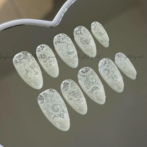 May include: A collection of ten white, oval-shaped false nails featuring a detailed lace pattern. The nails are displayed on a reflective surface, highlighting the intricate lace design. The text "Ming Original" is visible on the surface.