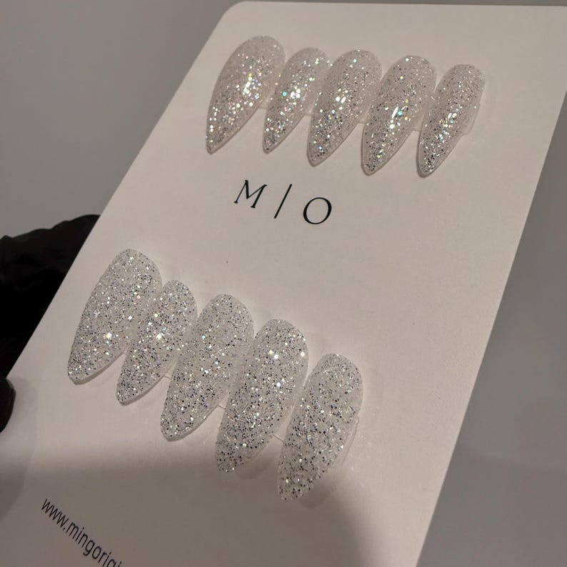 K&ouml;nnte beinhalten: Ein Set aus zehn mandelf&ouml;rmigen Kunstn&auml;geln, die mit Glitzer bedeckt sind. Die N&auml;gel haben eine klare Basis mit einer Beschichtung aus silbernem Glitzer. Die N&auml;gel sind auf einer wei&szlig;en Karte mit dem Text "M/O" angeordnet.