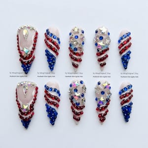 May include: A collection of ten artificial nails, each adorned with red, white, and blue embellishments. Some nails feature star-shaped gems, while others have a spiral design. The nails are decorated with rhinestones and gems.
