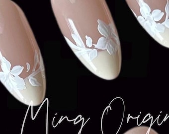 Può includere: Primo piano di quattro unghie ovali color nude con disegni floreali bianchi. Le unghie sono dipinte con uno smalto beige chiaro e hanno una punta bianca. I disegni floreali sono delicati e intricati, e sono disposti in uno schema simmetrico. Il testo "Ming Origin" è visibile nella parte inferiore dell'immagine.