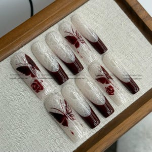 Pu&ograve; includere: Set di dieci unghie finte lunghe a forma di bara. Ogni unghia presenta un design in pizzo bianco, una punta bordeaux scuro e un accento di farfalla o rosa bordeaux. Le unghie sono disposte su una superficie chiara.
