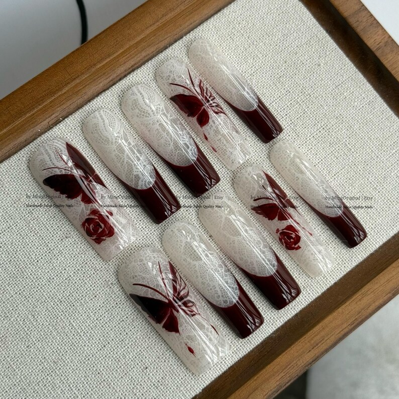 Pu&ograve; includere: Set di dieci unghie finte lunghe a forma di bara. Ogni unghia presenta un design in pizzo bianco, una punta bordeaux scuro e un accento di farfalla o rosa bordeaux. Le unghie sono disposte su una superficie chiara.