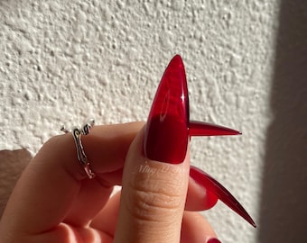 May include: Close-up of a hand with long, sharp, red stiletto nails. The nails are a glossy, opaque red.