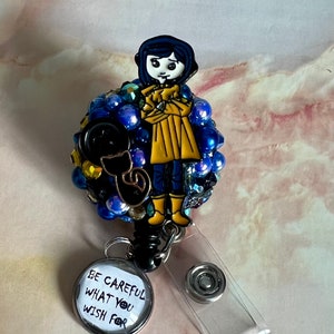 K&ouml;nnte beinhalten: Eine blaue und gelbe Comicfigur mit einer schwarzen Katze auf einem Bett aus blauen und goldenen Perlen. Die Figur tr&auml;gt einen gelben Regenmantel und hat schwarze Haare. Der Text auf dem Abzeichen lautet "Sei vorsichtig, was du dir w&uuml;nschst".