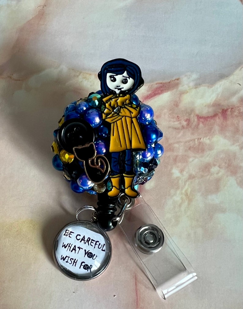 K&ouml;nnte beinhalten: Eine blaue und gelbe Comicfigur mit einer schwarzen Katze auf einem Bett aus blauen und goldenen Perlen. Die Figur tr&auml;gt einen gelben Regenmantel und hat schwarze Haare. Der Text auf dem Abzeichen lautet "Sei vorsichtig, was du dir w&uuml;nschst".