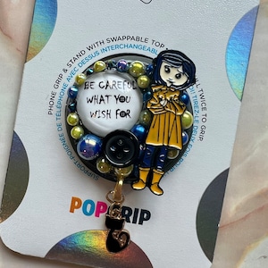 K&ouml;nnte beinhalten: Ein schwarzer und gelber Handy-Griff mit einer Comicfigur, die einen gelben Regenmantel und blaue Stiefel tr&auml;gt. Der Griff hat einen runden, wei&szlig;en Knopf mit dem Text "Sei vorsichtig, was du dir w&uuml;nschst". Der Griff ist an einem schwarzen Katzenanh&auml;nger befestigt.