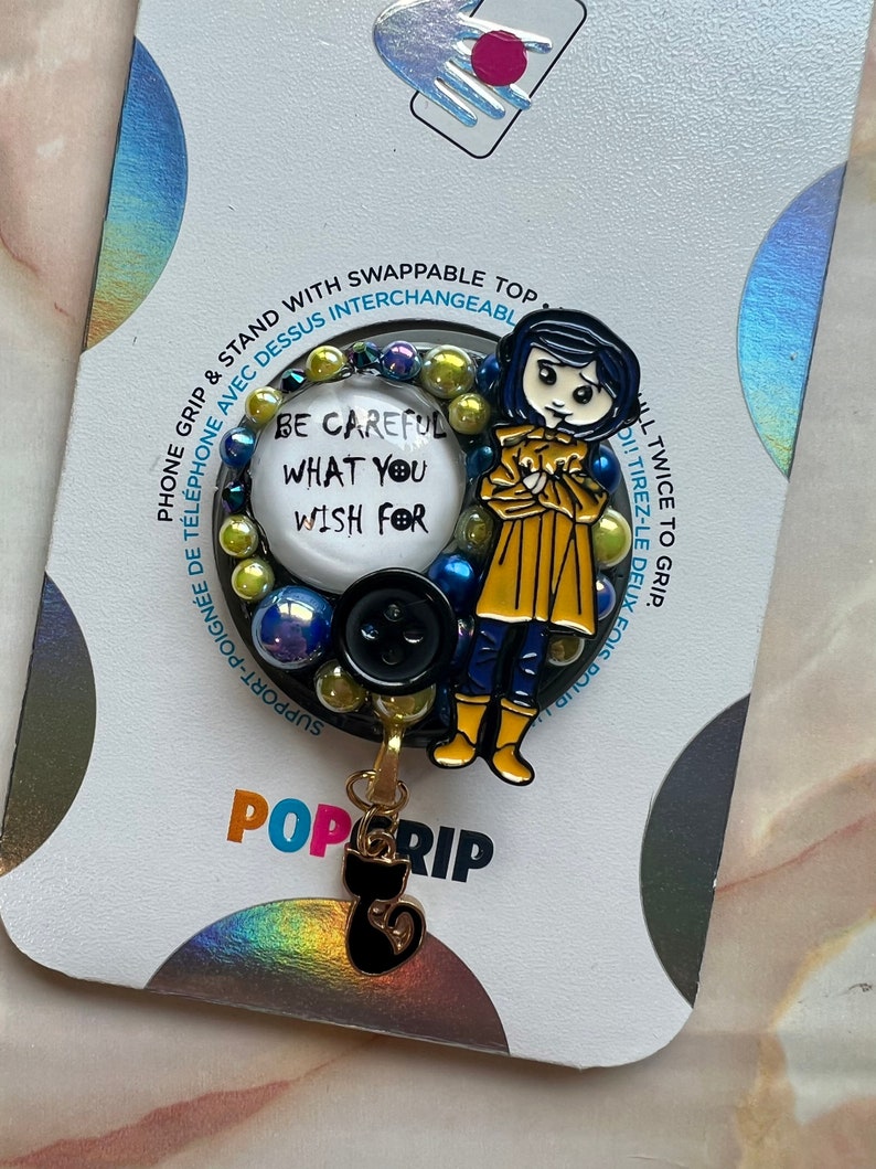 K&ouml;nnte beinhalten: Ein schwarzer und gelber Handy-Griff mit einer Comicfigur, die einen gelben Regenmantel und blaue Stiefel tr&auml;gt. Der Griff hat einen runden, wei&szlig;en Knopf mit dem Text "Sei vorsichtig, was du dir w&uuml;nschst". Der Griff ist an einem schwarzen Katzenanh&auml;nger befestigt.