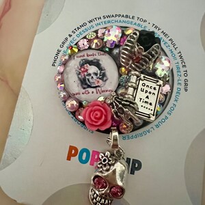 May include: A pink and silver phone grip with a skull charm. The grip has a removable top with a skull and flowers design and the text "I read books that end with a warning". The bottom of the grip has a book charm with the text "Once Upon A Time..."