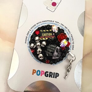 Op de afbeelding: Een zwarte telefoon grip met een schedel en kruisbeenderen ontwerp. De grip heeft een rood en zwart "Let op: Heet Content" bord en een zilveren bedel in de vorm van een pistool. De grip is bevestigd aan een witte achtergrond met het woord "POPGRIP" erop gedrukt.