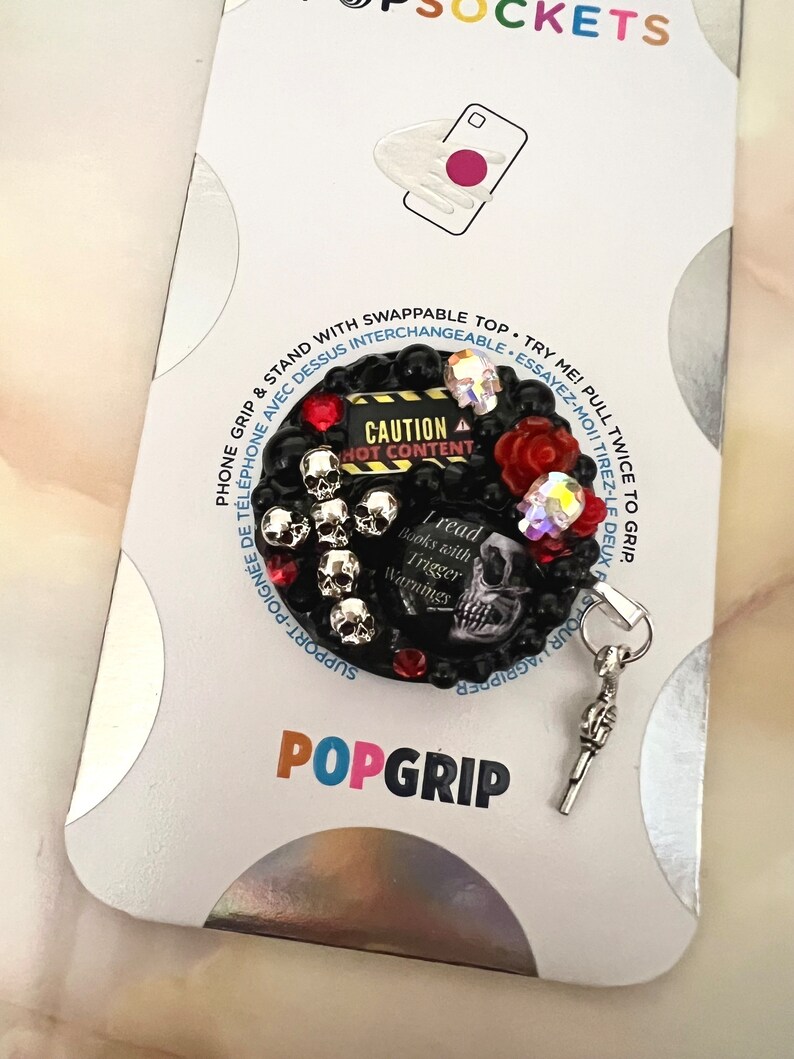 Op de afbeelding: Een zwarte telefoon grip met een schedel en kruisbeenderen ontwerp. De grip heeft een rood en zwart "Let op: Heet Content" bord en een zilveren bedel in de vorm van een pistool. De grip is bevestigd aan een witte achtergrond met het woord "POPGRIP" erop gedrukt.