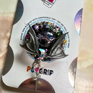 May include: A black and silver phone grip with a mountain scene and iridescent accents. The grip has a removable top and says "PopGrip" on the bottom.