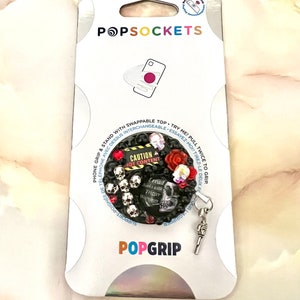 Op de afbeelding: Een zwarte en zilveren PopGrip-telefoonhouder met een schedel- en kruisbeenderontwerp. De grip heeft een rood hart, een rode roos en de tekst "CAUTION HOT CONTENT". De grip is bevestigd aan een zilveren sleutelhanger.