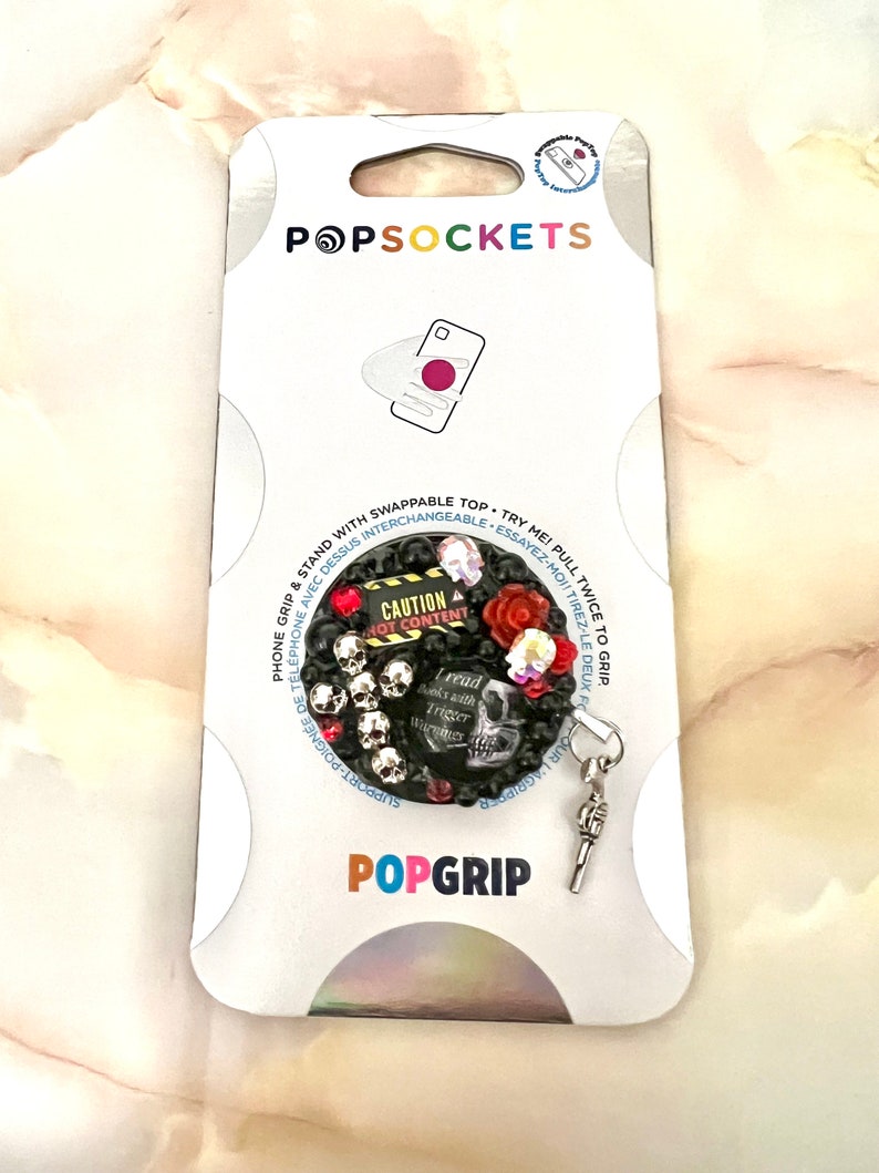 Op de afbeelding: Een zwarte en zilveren PopGrip-telefoonhouder met een schedel- en kruisbeenderontwerp. De grip heeft een rood hart, een rode roos en de tekst "CAUTION HOT CONTENT". De grip is bevestigd aan een zilveren sleutelhanger.