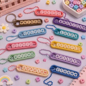 Può includere: Una collezione di portachiavi in plastica colorata con catenelle a palline, ciascuno contenente perline quadrate con lettere che compongono nomi come Cherry, Nika, Monica, Calvi, Luke, Olivia, Jessie, Mason, Hailey, Sophia ed Ethan, con perline a forma di cuore e stella.