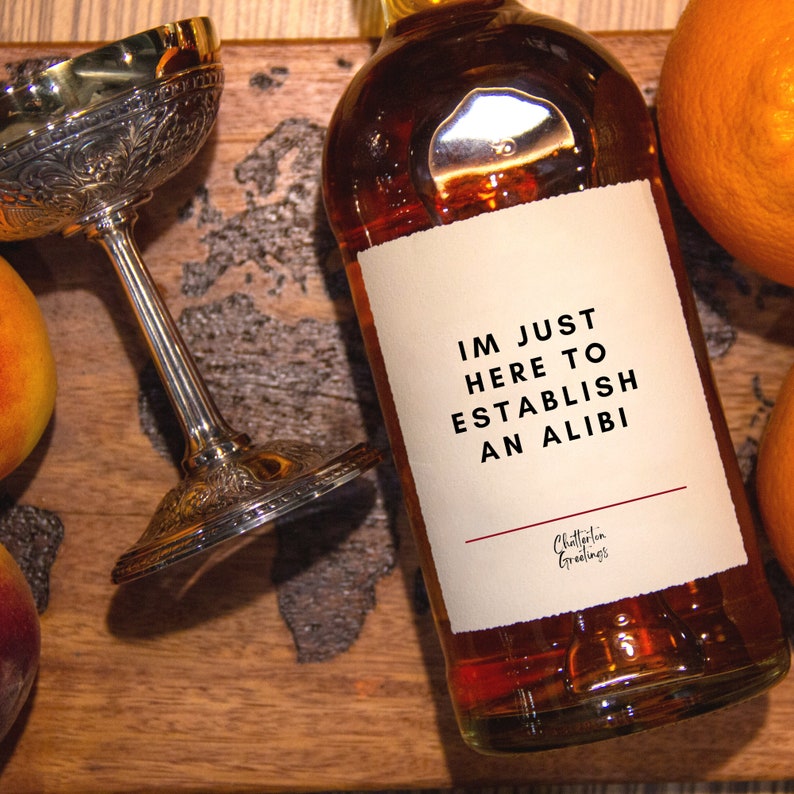 May include: A bottle of amber liquid with a white label that reads "IM JUST HERE TO ESTABLISH AN ALIBI." A silver goblet and fruit are on a wooden surface.
