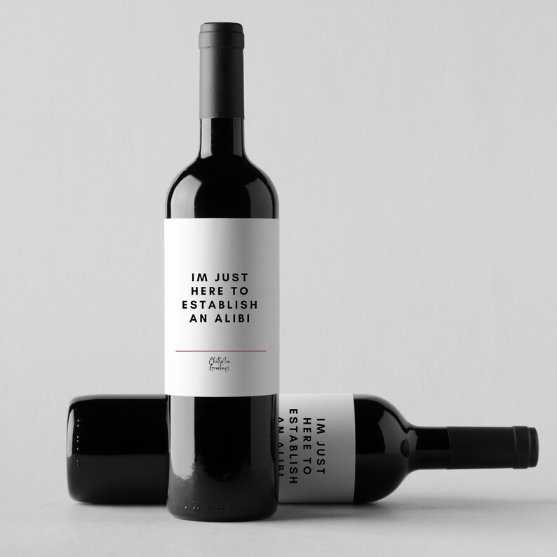 May include: Two black wine bottles with white labels. The labels read "IM JUST HERE TO ESTABLISH AN ALIBI" in black text. One bottle is upright, and the other is lying down. The bottles are on a white background.