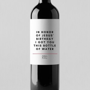 May include: A white wine bottle label with black text that reads "In honour of Jesus' birthday I got you this bottle of water". The label also includes the text "Chatterbox Greetings" in a smaller font.