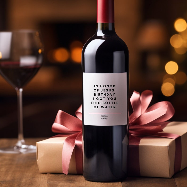 May include: A bottle of red wine with a white label that reads "In honour of Jesus' birthday I got you this bottle of water". The bottle is wrapped in a pink ribbon and a brown gift box.