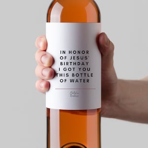 May include: A white wine bottle with a label that reads "IN HONOR OF JESUS' BIRTHDAY I GOT YOU THIS BOTTLE OF WATER". The label has a red line at the bottom and the text "Chatterton Greetings" in a smaller font.