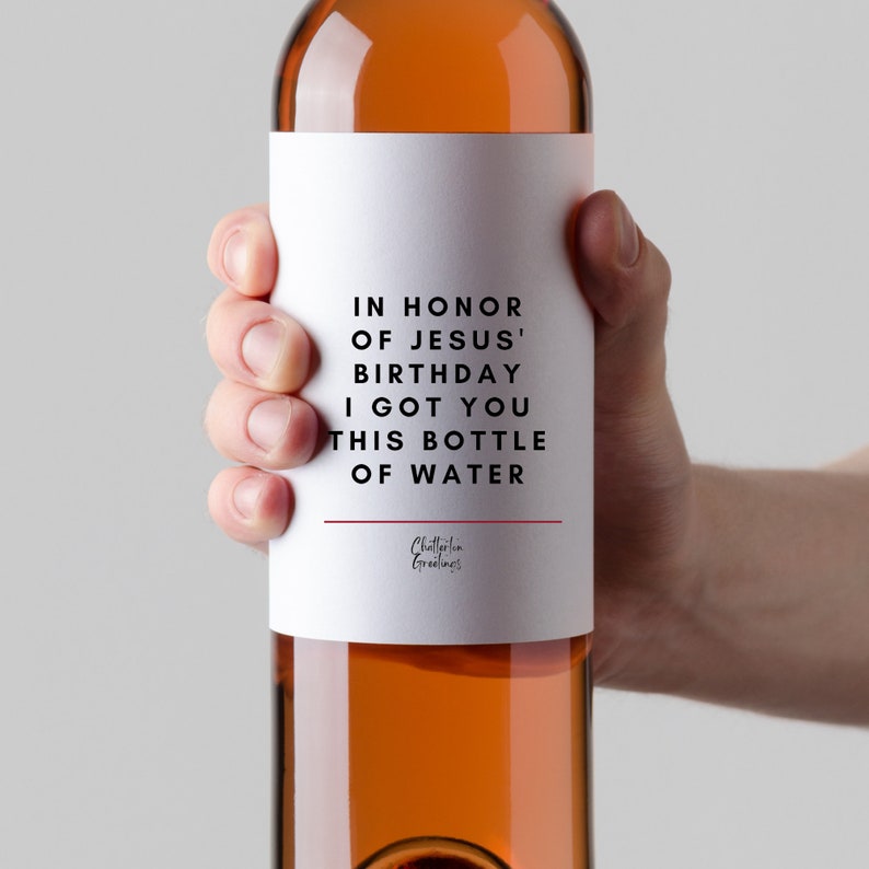 May include: A white wine bottle with a label that reads "IN HONOR OF JESUS' BIRTHDAY I GOT YOU THIS BOTTLE OF WATER". The label has a red line at the bottom and the text "Chatterton Greetings" in a smaller font.