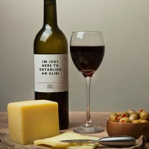 May include: A wine bottle with a label that reads "I'M JUST HERE TO ESTABLISH AN ALIBI" sits next to a wine glass filled with red wine. A block of yellow cheese, sliced cheese, and a bowl of olives complete the scene.