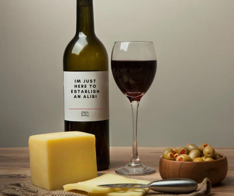 May include: A wine bottle with a label that reads "I'M JUST HERE TO ESTABLISH AN ALIBI" sits next to a wine glass filled with red wine. A block of yellow cheese, sliced cheese, and a bowl of olives complete the scene.