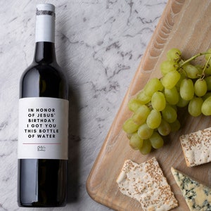 May include: A bottle of red wine with a white label that reads "In honour of Jesus' birthday I got you this bottle of water". The bottle is on a white marble surface with a wooden chopping board in the background. The chopping board has a bunch of green grapes, a wedge of blue cheese, and two pieces of flatbread with sesame seeds.
